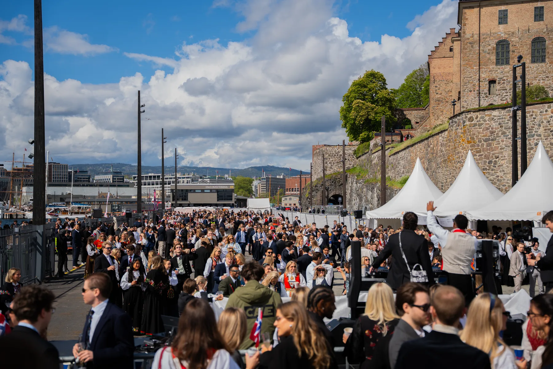 Bred oversikt over publikum og festivalområdet på Nasjonalfesten, med Akershus festning og Oslofjorden i bakgrunnen.